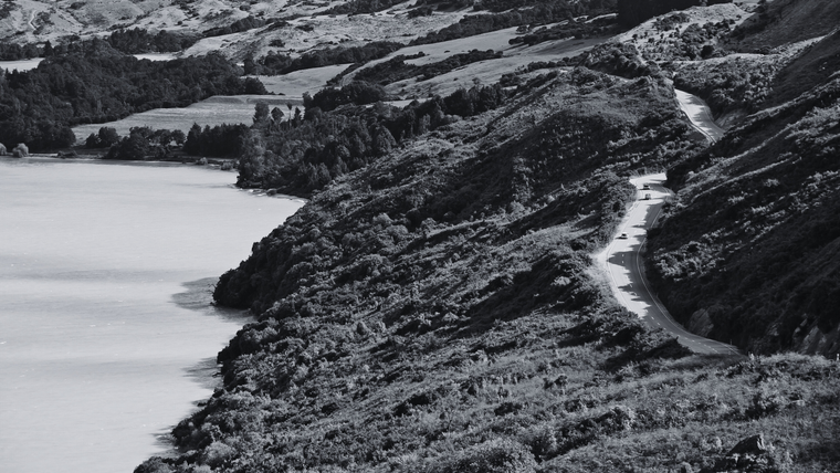 A winding coastal road in New Zealand, cutting through lush hills alongside a serene body of water