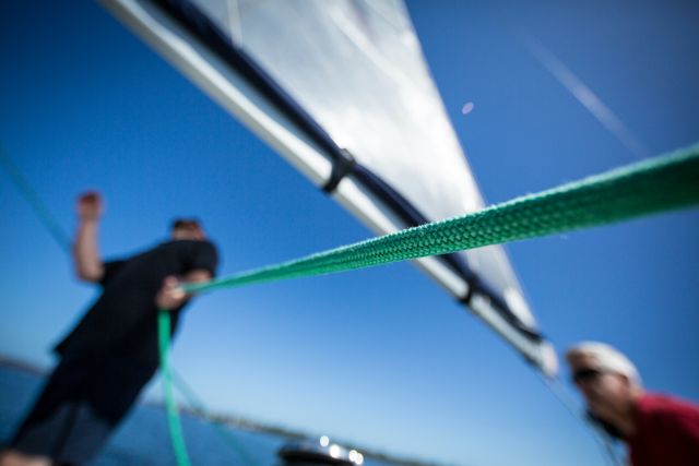 Close-up photo of a green rope on a sailboat, with blurred figures in the background and a sail catching the wind against a clear blue sky.
