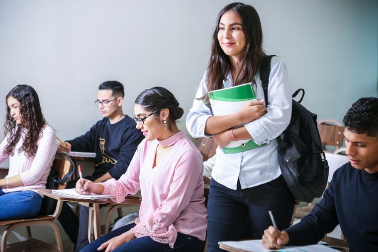 A group of college students sit in a classroom taking notes, while one student stands beside her desk holding notebooks and a backpack.
